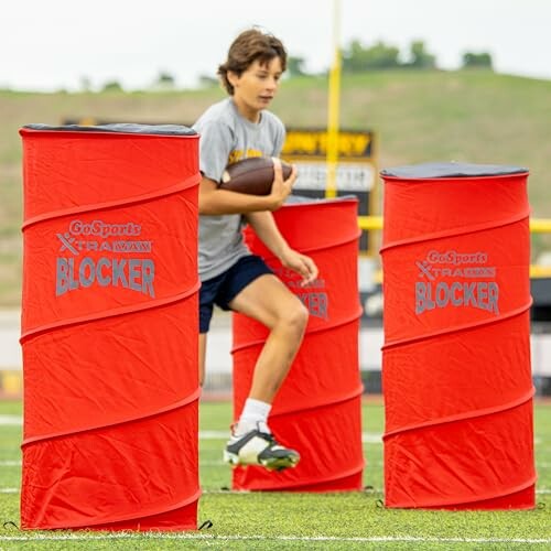 A person holding a football running through red training blockers on a field.