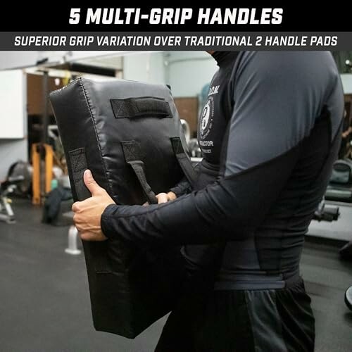 Person holding a black exercise pad with multiple grip handles in a gym.