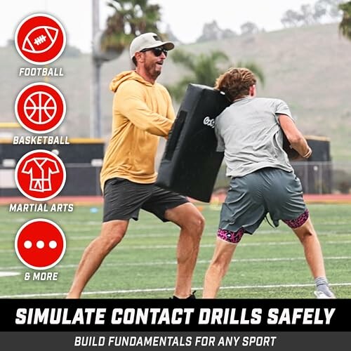 Two people practicing contact drills with a padded shield on a sports field.