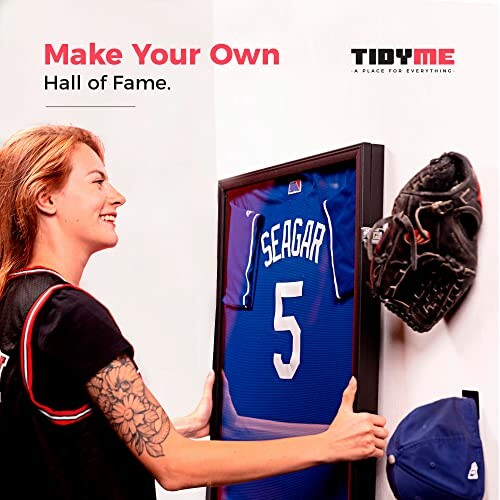 Person hanging a framed baseball jersey on a wall with a glove and cap nearby.